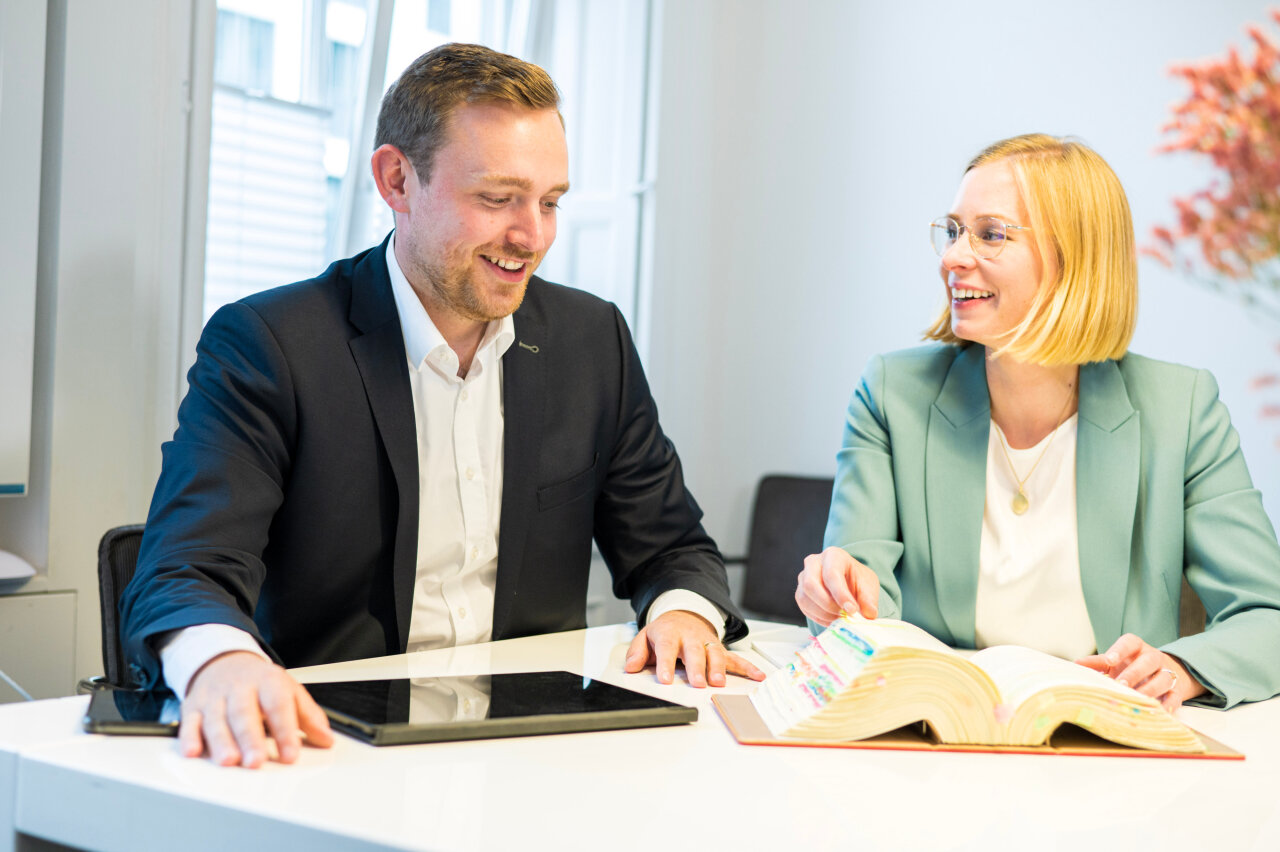 Zwei EGIDO-Mitarbeiter sitzen an einem Tisch in einem modernen Büro. Der Mann trägt einen dunklen Anzug und lächelt, während die Frau in einem hellen Blazer eine offene Buchseite zeigt und ebenfalls lächelt. Auf dem Tisch liegen ein Tablet und ein geöffnetes Buch mit bunten Markierungen. In der Hintergrund ist eine helle Wand und Pflanzen zu sehen.