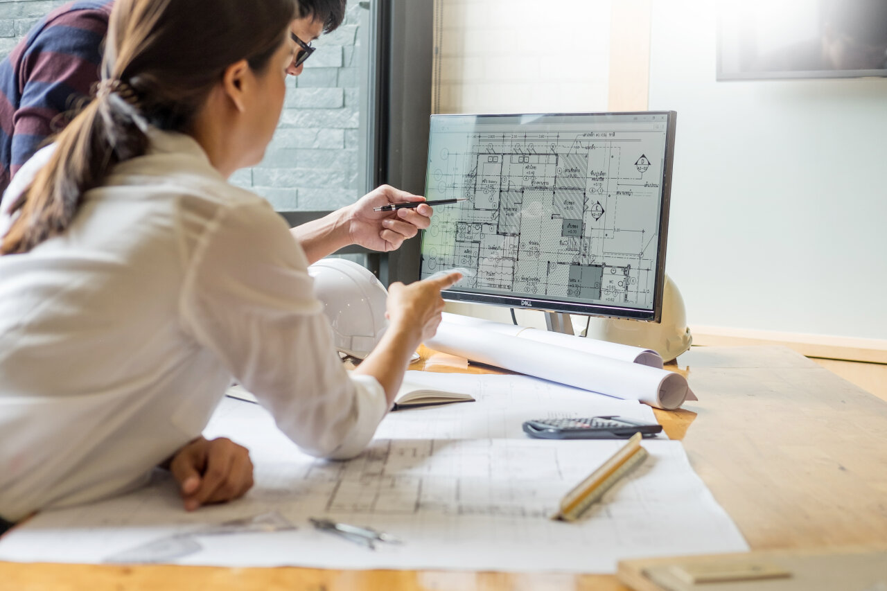 Zwei Architekten und Bauingenieure betrachten einen Bauplan auf einem Computerbildschirm in einem modernen Büroumfeld. Auf dem Tisch liegen Zeichnungen und ein Architekturhelm, während eine Person mit einem Stift auf den Bildschirm deutet.